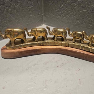 May include: A decorative brass elephant parade sculpture. Five golden elephants are arranged in a row on a brass bridge, mounted on a curved wooden base. The elephants are detailed with textured skin and raised trunks.