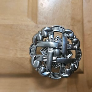 Może przedstawiać: Srebrny uchwyt do szafki o skomplikowanym wzorze. Uchwyt ma centralny geometryczny wzór otoczony okrągłą ramą ze stylizowanymi motywami dłoni. Tło to jasne drewniane drzwi szafki.