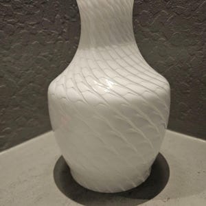 May include: A white glass vase with a narrow neck and a rounded body, featuring a subtle, repeating diagonal wave pattern across its surface. The glass has a semi-translucent quality, creating a soft texture that wraps around the entire vessel.