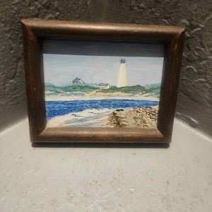 Puede incluir: Pintura enmarcada de una escena costera. La obra representa un faro blanco, pequeñas casas y un océano azul con olas blancas. El marco es de madera marrón oscuro.
