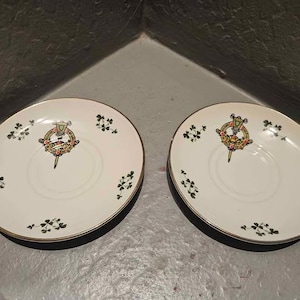 May include: Two white saucers with a gold rim and a black base. Each saucer features a central emblem with red, green, and gold details, surrounded by small green shamrock designs. The saucers are placed side by side.