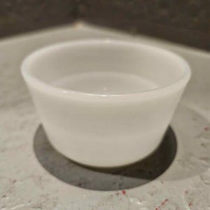 Könnte beinhalten: Eine kleine, runde, weiße Keramikschale mit glatten, konisch zulaufenden Seiten und einer weiten Öffnung. Die Schale hat ein schlichtes Design mit einer glänzenden Oberfläche, die das Licht gleichmäßig auf der gewölbten Form reflektiert.