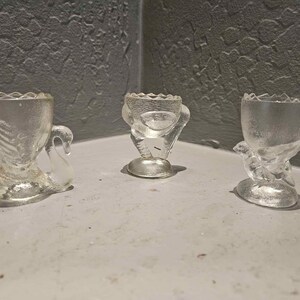 May include: Three clear glass egg cups with textured surfaces. The left cup features a swan-shaped base, the center cup has a bird-like figure with legs, and the right cup is shaped like a small bird perched on a base. Each cup has a scalloped rim.