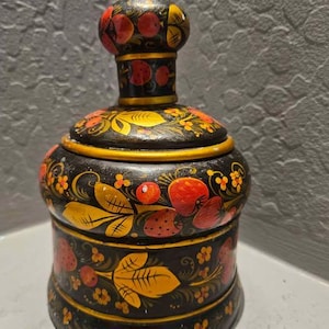 May include: A hand-painted, wooden trinket box with a lid. The box is black with a gold rim and is decorated with red strawberries, yellow leaves, and small orange flowers. The lid has a rounded top with a knob.