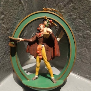 May include: A decorative ornament featuring a figure in a brown coat and yellow trousers carrying a smaller figure on their shoulders. The figures are framed by a green circular border with a gold rim and a gold hanging cord attached at the top.