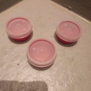 Puede incluir: Tres pequeños recipientes redondos rojos con tapas rosas. Las tapas son translúcidas con un borde blanco y un diseño circular central. Los recipientes están dispuestos sobre una superficie clara y texturizada.