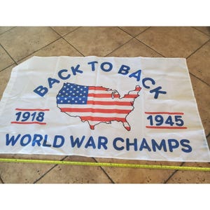 Puede incluir: Bandera blanca con el texto azul "BACK TO BACK WORLD WAR CHAMPS". La bandera presenta un mapa de los Estados Unidos con el diseño de la bandera estadounidense. Los años 1918 y 1945 están impresos en rojo.