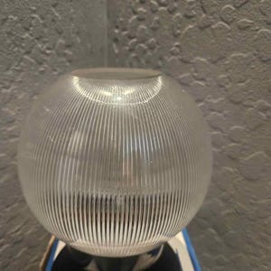 May include: A spherical glass light fixture shade featuring a clear, ribbed texture with vertical lines running from the top to the bottom. The globe has a circular opening at the top and a wider base, designed to diffuse light through the glass.