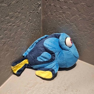 Può includere: Un peluche a forma di pesce chirurgo blu con un corpo in tessuto morbido e testurizzato. Il pesce presenta una testa e un corpo blu brillante, strisce blu scuro e vivaci accenti gialli sulla coda e sulla pinna pettorale, con un grande occhio bianco.