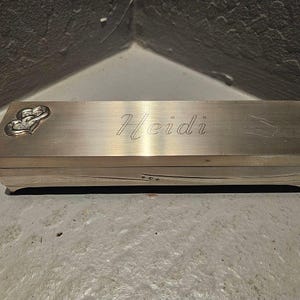 Könnte beinhalten: Eine rechteckige Box aus gebürstetem Metall mit dem eingravierten Namen "Heidi" auf der Oberseite. Zwei herzförmige Verzierungen mit Diamantakzenten befinden sich auf der linken Seite. Die Box hat ein schlichtes, elegantes Design.