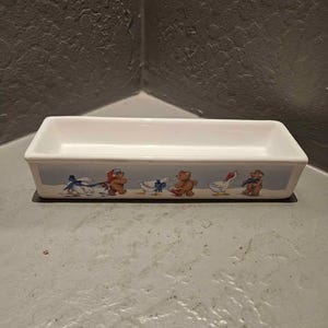 May include: A rectangular white ceramic loaf pan featuring a decorative band along the exterior side. The design displays a series of illustrated brown teddy bears and white geese wearing blue and red scarves and hats against a light blue background.