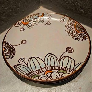 Op de afbeelding: Een decoratieve keramische plaat met een crèmekleurig oppervlak en een bruine rand. De plaat heeft handgeschilderde bloemen en abstracte ontwerpen in bruin, blauw, oranje en groen. De plaat is rond en lijkt gestapeld met een andere plaat.