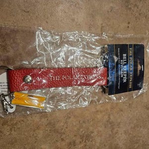 May include: A red textured leather keychain with a silver snap button and the text THE POLAR EXPRESS embossed in gold. It includes a small yellow rectangular tag and a dark blue cardboard header card labeled THE POLAR EXPRESS BELL KEEPER.