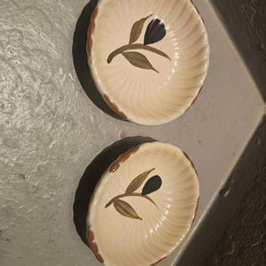 May include: Two cream-coloured ceramic dishes with a scalloped edge and a painted floral design. Each dish features an olive-shaped design in brown, green, and black. The dishes are likely for serving or display, possibly for olives.