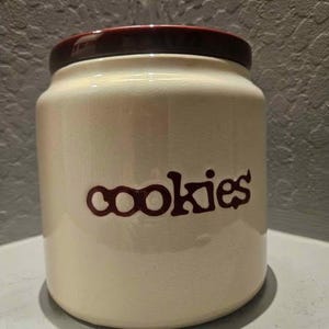 Pode incluir: Um pote cilíndrico de cerâmica para biscoitos com corpo na cor creme e acabamento em esmalte craquelado. O pote possui uma tampa marrom escura e a palavra cookies está impressa em marrom escuro com uma fonte estilizada em minúsculas.