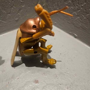 Puede incluir: Un juguete de plástico detallado que representa un insecto, con una cabeza de color cobre, un cuerpo amarillo y patas beige. El juguete tiene extremidades y antenas articuladas, y está posicionado de forma dinámica. Probablemente forma parte de una colección de juguetes de insectos.