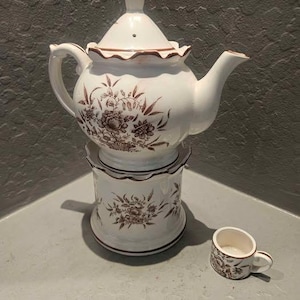 May include: A white ceramic teapot with a lid and a matching warmer base, both decorated with brown floral patterns and scalloped edges. A small, matching white ceramic teacup with a brown floral design sits beside the teapot and warmer.
