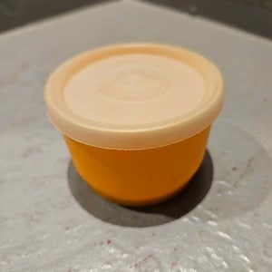 May include: A small, round, orange storage container with a matching light peach-colored circular lid. The container has a smooth, matte finish and a simple, functional design, featuring a slightly tapered base and a secure, fitted top cover.