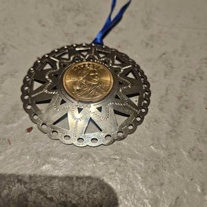 May include: A circular silver-toned metal ornament featuring a cut-out star pattern and a scalloped edge. A gold-colored 2000 Liberty Sacagawea dollar coin is set in the center. A blue ribbon is attached to the top for hanging the decorative piece.