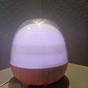 Peut inclure: Un diffuseur d'huiles essentielles en forme d'œuf avec une base en imitation bois clair et un dessus transparent en forme de dôme. Le diffuseur émet une douce lumière blanche. Trois boutons de commande sont visibles à l'avant de la base.