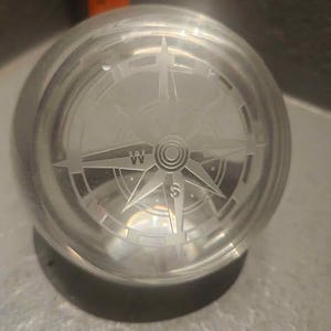 Puede incluir: Un pisapapeles de cristal transparente con un diseño de rosa de los vientos grabado en su interior. La rosa de los vientos presenta indicadores direccionales y un diseño circular central. El pisapapeles es redondo.