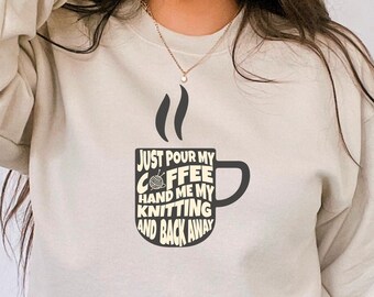 May include: A light beige sweatshirt with a black graphic of a coffee mug that says "Just pour my coffee hand me my knitting and back away".