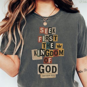 May include: A dark grey t-shirt featuring the text "SEEK FIRST THE KINGDOM OF GOD" in a collage style. The text is in various colours and fonts. The shirt also has the text "MATTHEW 6:33" at the bottom.