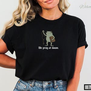 May include: A black t-shirt featuring a graphic of a light green frog holding a brown book and a rosary. Below the frog, the text reads, We pray at dawn. The shirt has short sleeves and a crew neckline, providing a simple and direct design.