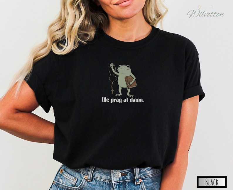 May include: A black t-shirt featuring a graphic of a light green frog holding a brown book and a rosary. Below the frog, the text reads, We pray at dawn. The shirt has short sleeves and a crew neckline, providing a simple and direct design.