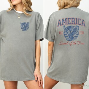 Puede incluir: Una camiseta gris con un gr&aacute;fico de estilo vintage de un &aacute;guila sosteniendo una pancarta con las palabras "AMERICA EST 1776 Land of the Free".