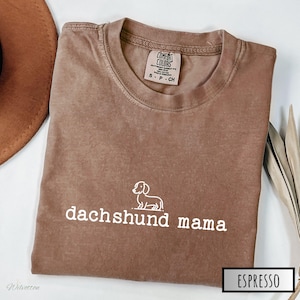 May include: A brown t-shirt with the text "dachshund mama" and a small outline of a dachshund dog. The shirt is made of a soft, comfortable fabric and has a distressed look. The text is in white and the dog is in white. The shirt is folded and laid flat on a white surface.