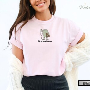 May include: A light pink t-shirt featuring a graphic of a green frog holding a rosary and a book, with the text "We pray at dawn." printed in black below the illustration. The shirt has a crew neckline and short sleeves, worn with a white cardigan.