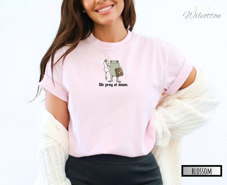 May include: A light pink t-shirt featuring a graphic of a green frog holding a rosary and a book, with the text "We pray at dawn." printed in black below the illustration. The shirt has a crew neckline and short sleeves, worn with a white cardigan.