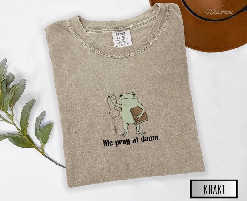 May include: A khaki-colored crew neck t-shirt featuring a graphic of a green frog holding a brown book and a rosary. Below the frog, the text reads "We pray at dawn." The shirt has a Comfort Colors brand label at the inner neckline.