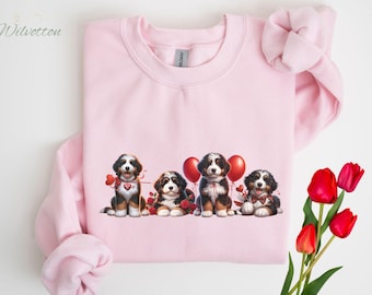 May include: Pink sweatshirt with a graphic of four Bernese Mountain Dog puppies holding hearts and balloons. The puppies are wearing red collars and have black and white fur.