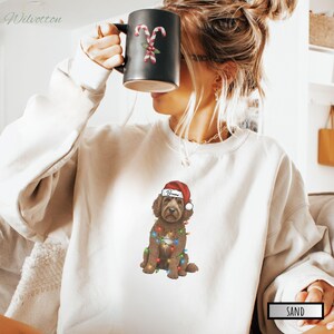 May include: A sand-colored sweatshirt featuring a brown dog wearing a Santa hat and Christmas lights. A black mug with candy cane designs is held up. The word "Bears" is on the hat. The word "Sand" is in a rectangle.