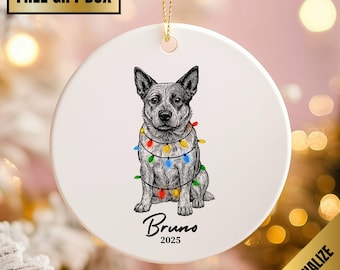 May include: A white ceramic Christmas ornament featuring a detailed illustration of a dog adorned with colorful string lights. The dog's name, "Bruno," and the year "2025" are written below the dog.