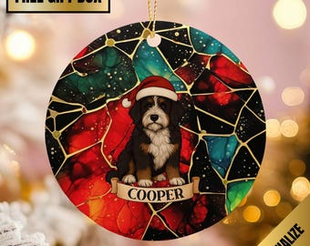 May include: Round Christmas ornament featuring a Bernedoodle puppy wearing a Santa hat. The ornament has a stained glass design in red, green, and blue with gold accents. The puppy's name, "COOPER," is on a banner.