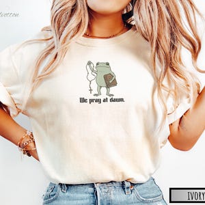 May include: An ivory short-sleeved t-shirt featuring a graphic of a green frog holding a rosary and a brown book. Below the frog, the black text reads, "We pray at dawn." The shirt has a crew neckline and a relaxed fit, showcasing a simple design.