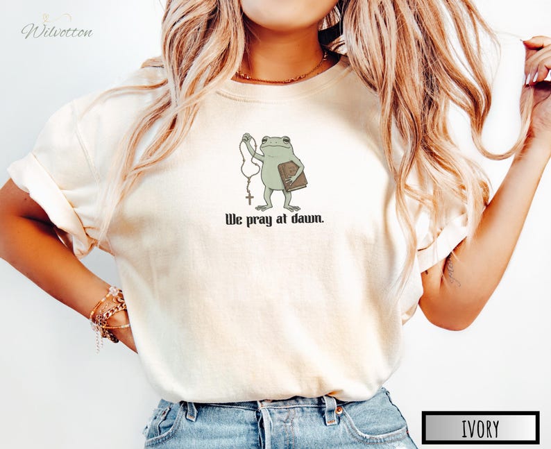 May include: An ivory short-sleeved t-shirt featuring a graphic of a green frog holding a rosary and a brown book. Below the frog, the black text reads, "We pray at dawn." The shirt has a crew neckline and a relaxed fit, showcasing a simple design.