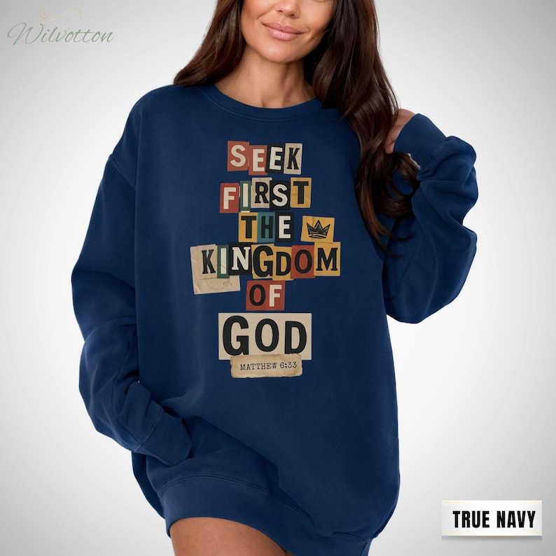 May include: A navy blue sweatshirt featuring the text "SEEK FIRST THE KINGDOM OF GOD" in a collage design. The text is in various colours and fonts. The sweatshirt also has the text "MATTHEW 6:33" at the bottom.