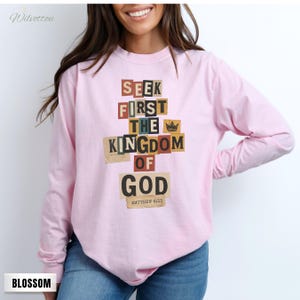 May include: A pale pink long-sleeved top featuring the phrase "SEEK FIRST THE KINGDOM OF GOD" in a collage design. The text is in various colours and fonts. The top also includes the text "MATTHEW 6:33".