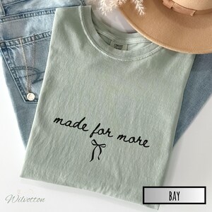 Puede incluir: Una camiseta verde claro con las palabras "made for more" en escritura negra y un lazo. La camiseta est&aacute; doblada sobre unos vaqueros azules, con un collar dorado y un sombrero beige.