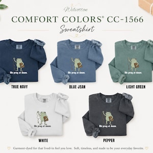 May include: Five Comfort Colors CC-1566 crewneck sweatshirts in true navy, blue jean, light green, white, and pepper. Each features a graphic of a frog holding a rosary and a book with the text "We pray at dawn." The sweatshirts have long sleeves.