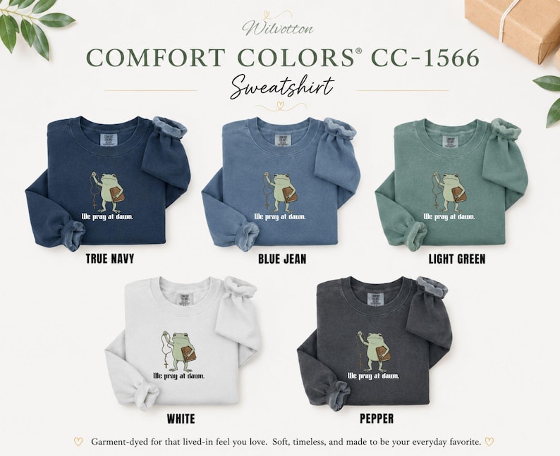 May include: Five Comfort Colors CC-1566 crewneck sweatshirts in true navy, blue jean, light green, white, and pepper. Each features a graphic of a frog holding a rosary and a book with the text "We pray at dawn." The sweatshirts have long sleeves.