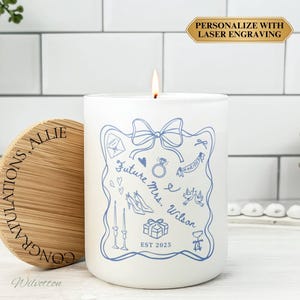 May include: A white candle with a blue illustration and the text "Future Mrs. Wilson EST 2025". The candle has a wooden lid with the text "CONGRATULATIONS, ALLIE". The candle is lit and is a wedding or engagement gift.