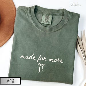 Puede incluir: Una camiseta verde musgo con las palabras "made for more" en escritura blanca y un gr&aacute;fico de lazo. La camiseta es de la marca Comfort Colors, talla S-P-CH. Un sombrero marr&oacute;n y hojas secas est&aacute;n en el fondo.