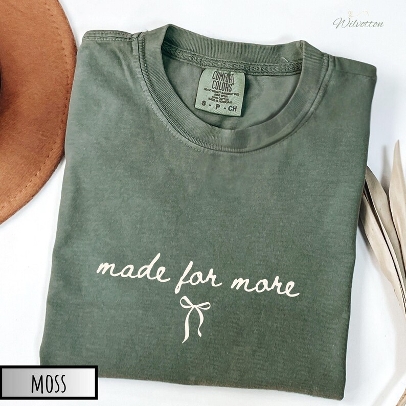 Puede incluir: Una camiseta verde musgo con las palabras "made for more" en escritura blanca y un gr&aacute;fico de lazo. La camiseta es de la marca Comfort Colors, talla S-P-CH. Un sombrero marr&oacute;n y hojas secas est&aacute;n en el fondo.