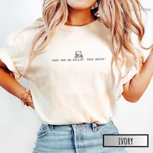 May include: A white t-shirt with a golf cart graphic and the text "They see me rollin' they hatin'". The shirt is shown on a model wearing blue jeans. The color of the shirt is ivory.
