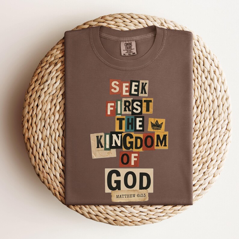 May include: A brown t-shirt featuring the text "SEEK FIRST THE KINGDOM OF GOD" in a collage design. The text uses different colours and fonts. The folded shirt rests on a circular woven mat.